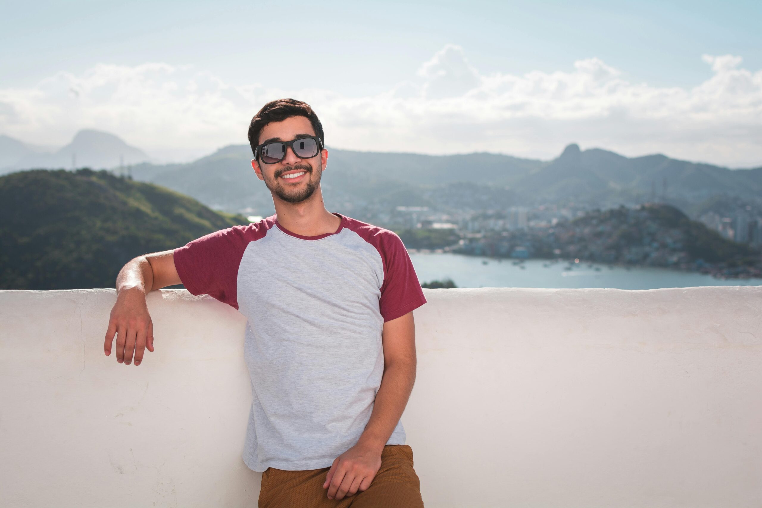 Home Young man in sunglasses leaning on a balcony with a scenic city view in the background.